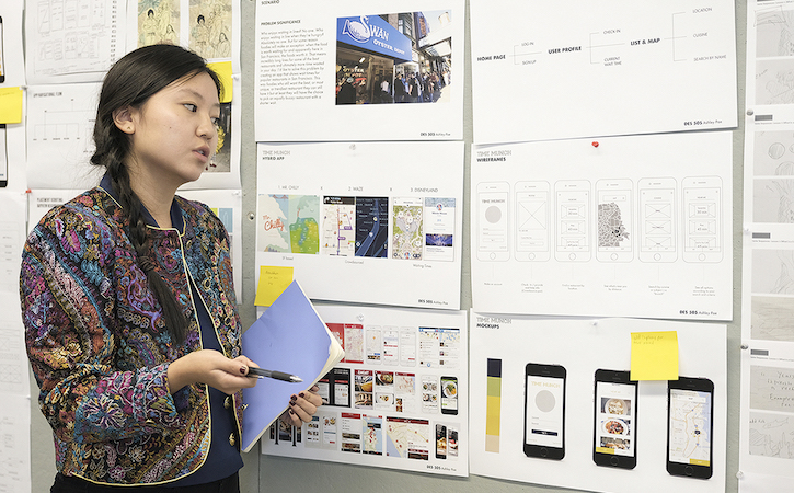 Instructor gesturing towards wall with design schematics on paper