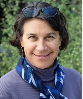 Close-up outdoor portrait of a person with short dark hair, wearing a purple turtleneck sweater and a blue patterned scarf tied around the neck. A pair of dark sunglasses rests on top of the head. The person is smiling and wearing small dangling earrings. The background is softly blurred green foliage, giving a natural, garden-like setting.