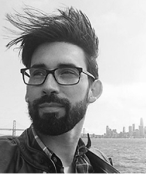 Black-and-white portrait of a person standing outdoors near a body of water, wearing rectangular eyeglasses, a dark jacket, and a collared shirt. The person has short facial hair and windblown hair swept to one side. In the background, a suspension bridge and a distant city skyline are visible under an overcast sky.